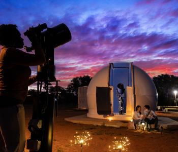sunset image with woman looking through telescope and couple sitting in front of dome