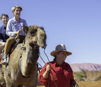 image of two people on camel being led by a guide with Uluru in background