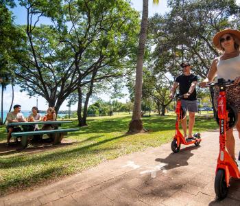 Man and woman on scooters riding along Darwin Esplanade. People enjoying picnic in the background.