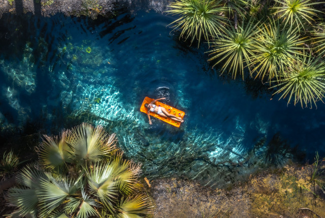 A woman floats on an orange lilo down Bitter Springs Thermal pool surrounded by palm trees