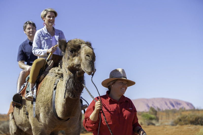 image of two people on camel being led by a guide with Uluru in background